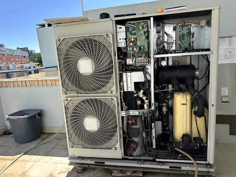 Unidade exterior aberta numa cobertura, com placas de comando, compressor e kit hidráulico à vista.