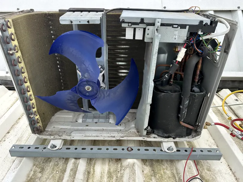 Unidade exterior desmontada num telhado de chapa, com o ventilador azul, o compressor e os manómetros ligados.
