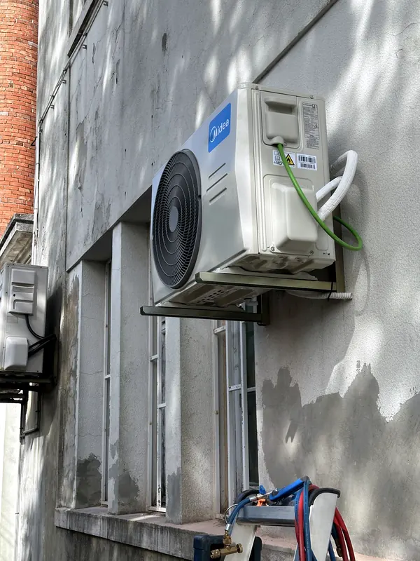 Unidade exterior Midea montada em consola numa parede rebocada, com o grupo manométrico e as mangueiras de carga no escadote.
