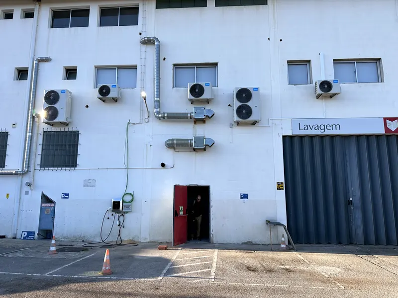 Cinco unidades exteriores e condutas de extração montadas junto às janelas do piso superior, na fachada de um edifício industrial.